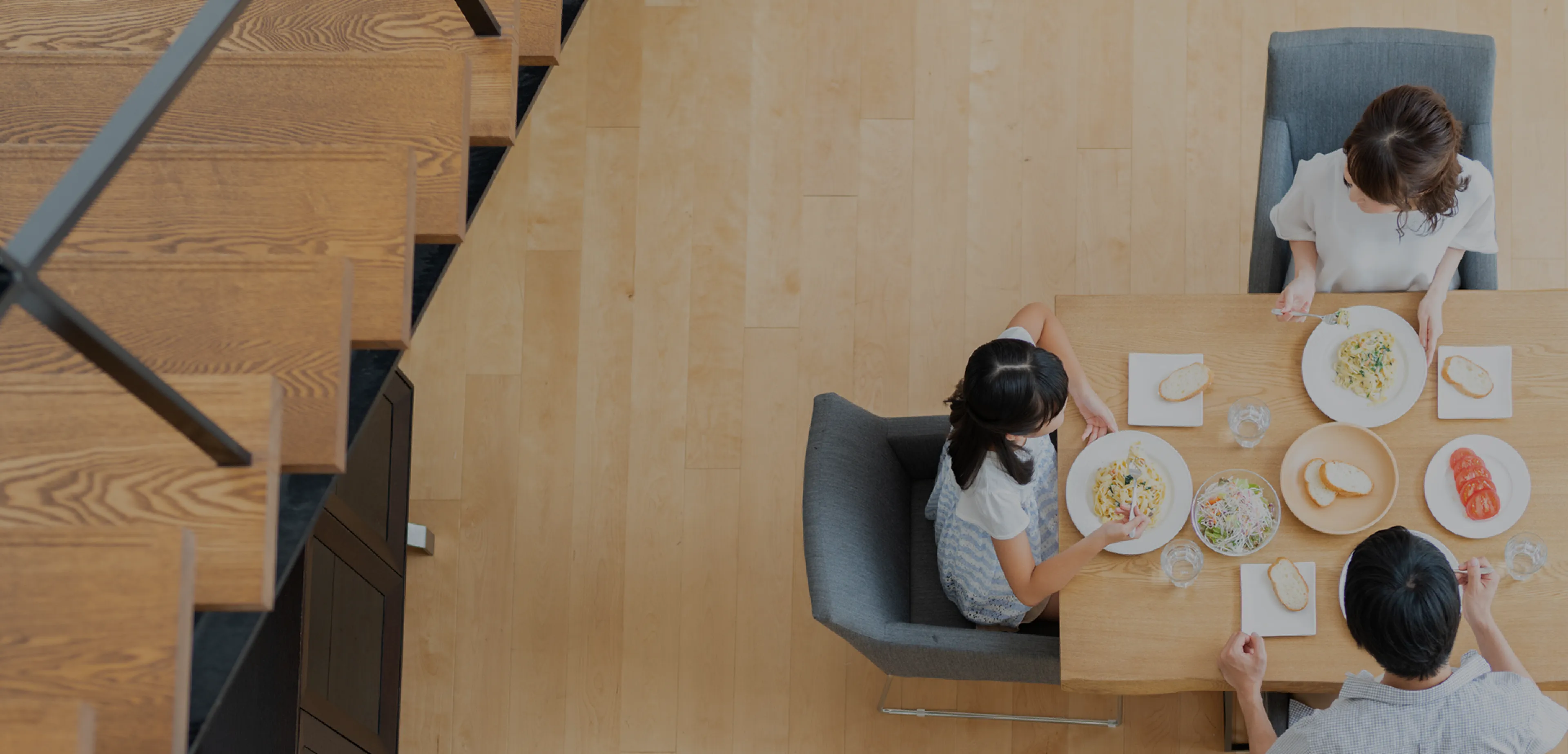 木の床のリビングとダイニングを上から見た様子。ローテーブルとベージュの椅子、マグを手に歩く女性、くつろぐ人の足元が写る明るい住空間